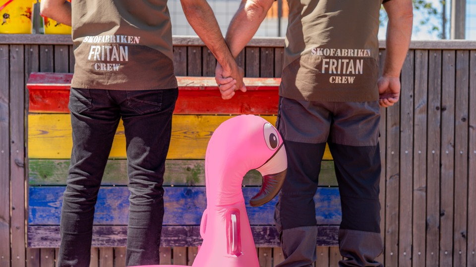 Två killar håller varandra i handen framför en regnbågsfärgad vägg.