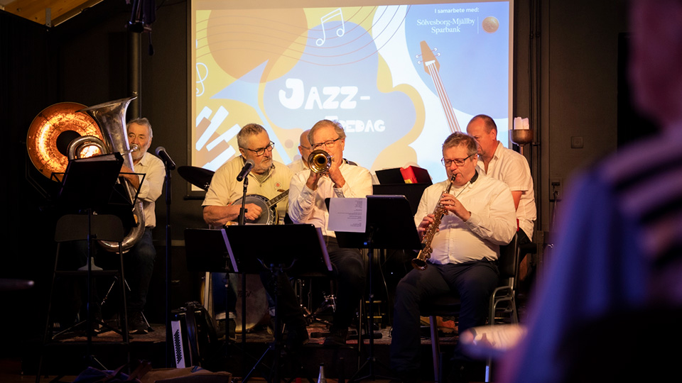 Ett jazzband sitter på scen och spelar olika blåsinstrument, klädda i vit skjorta.