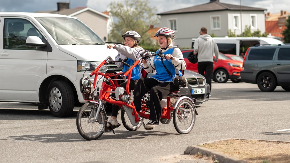 Två tjejer cyklar på en Paracykel på en parkering. De ser glada ut.