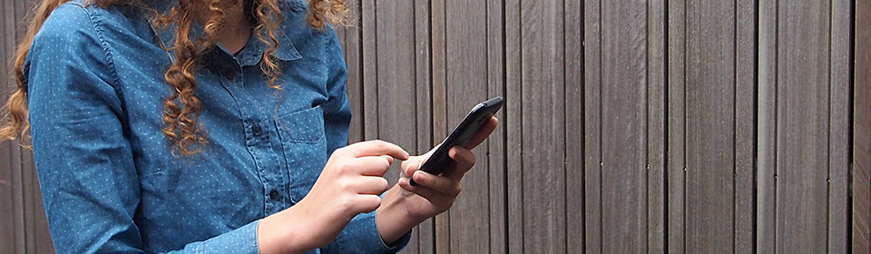 En ung tjej klädd i en blå skjorta och långt brunt lockigt hår håller en mobiltelefon i handen.