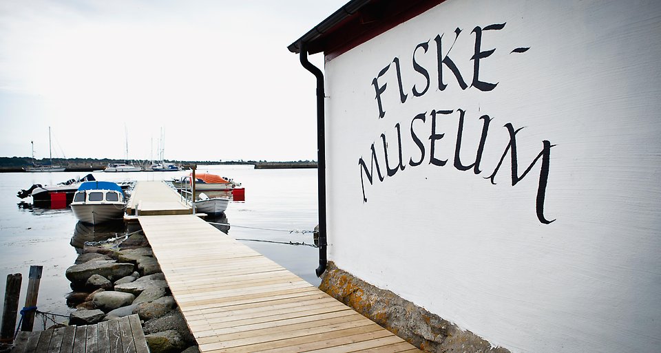 Utsikt över fiskemuseum i Hällevik. Det är en vit byggnad med texten Fiskemuseum. Havet och en brygga syns till vänster i bilden. 