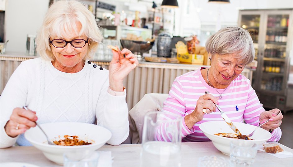 Två kvinnor som sitter bredvid varandra och äter. Den ena kvinnan har en vit tröja och den andra har en rosa tröja med vita ränder. 