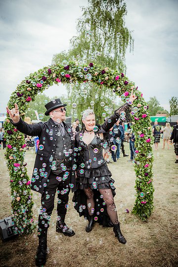 Par klädda i svart läder står tillsammans under blomsterbåge och det blåses ut såpbubblor över paret.