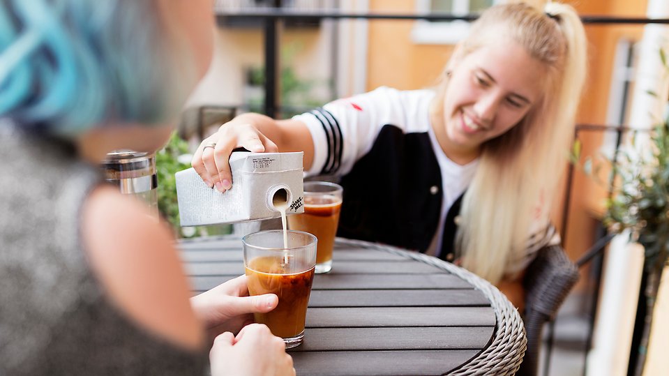 En kvinna med blont hår häller upp mjölk i ett kaffeglad åt en annan person.